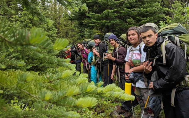 A line of people wearing backpacks pause hiking for a photo amid thick green trees.